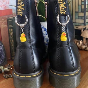 May include: A pair of black leather ankle boots featuring yellow stitching along the sole and a black heel pull tab with yellow text. Each boot is adorned with a silver-toned metal ring charm featuring a small yellow rubber duck pendant.