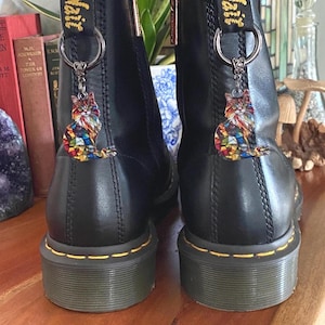 May include: A pair of black leather ankle boots featuring yellow stitching along the sole and a black heel loop with yellow AirWair text. Each boot is adorned with a silver-toned metal ring holding a colorful stained-glass style cat charm.