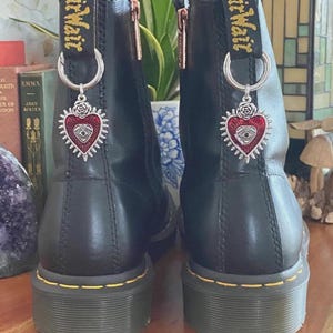 May include: A pair of black leather boots featuring yellow stitching along the sole and a black heel pull tab with yellow AirWair text. Each boot is adorned with a silver-toned metal charm shaped like a heart with a central eye and rose.