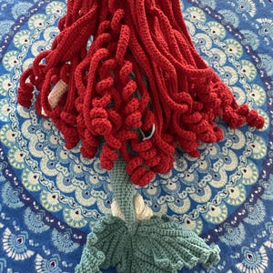 Pode incluir: Boneca de sereia de croch&ecirc; feita &agrave; m&atilde;o com cabelo vermelho vibrante, corpo azul claro e cauda verde-azulado. O cabelo da boneca &eacute; estilizado em mechas longas e encaracoladas. A cauda tem formato de leque. A boneca est&aacute; colocada sobre uma superf&iacute;cie azul estampada.