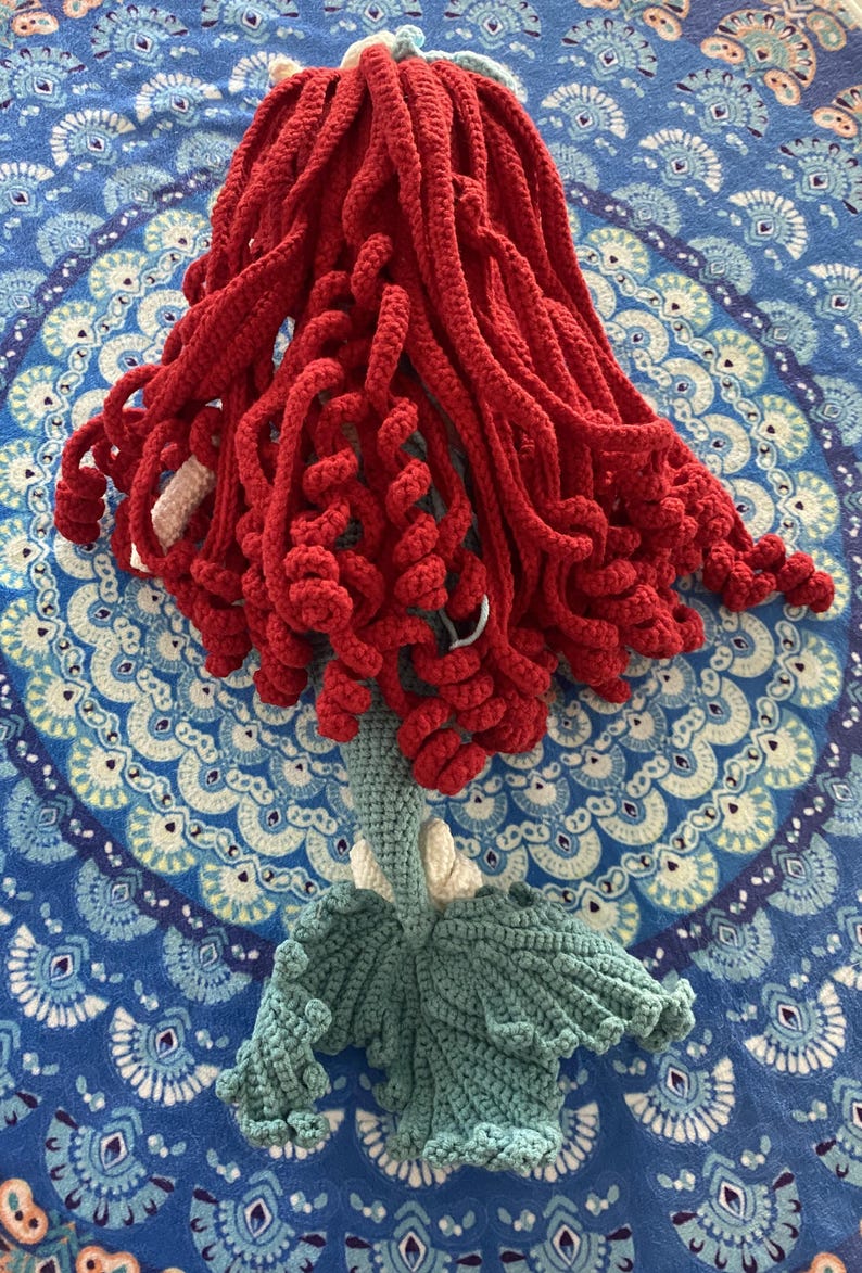 Pode incluir: Boneca de sereia de croch&ecirc; feita &agrave; m&atilde;o com cabelo vermelho vibrante, corpo azul claro e cauda verde-azulado. O cabelo da boneca &eacute; estilizado em mechas longas e encaracoladas. A cauda tem formato de leque. A boneca est&aacute; colocada sobre uma superf&iacute;cie azul estampada.