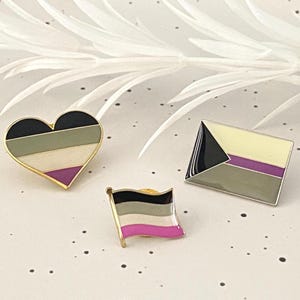 May include: Three enamel pins with gold borders. One is heart-shaped, the others are rectangular. Each pin features a design with black, green, white, gray, and purple colors. The pins are on a white surface with a decorative white leaf.
