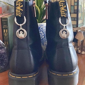 May include: A pair of black leather boots featuring yellow stitching along the thick soles and black heel loops with the yellow text AirWair. Each boot has a silver-toned circular charm depicting a phoenix attached to the heel loop by a ring.