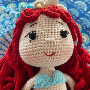 Pode incluir: Boneca de croch&ecirc; feita &agrave; m&atilde;o com cabelo vermelho, olhos pretos e um top azul claro. A boneca tem uma express&atilde;o sorridente e &eacute; adornada com uma tiara branca e azul claro. A boneca &eacute; feita de fio e provavelmente &eacute; uma personagem de um filme de anima&ccedil;&atilde;o.