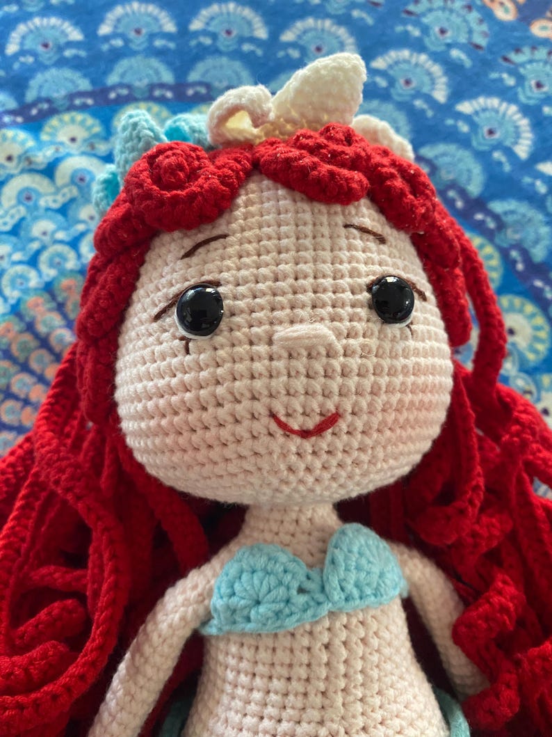 Pode incluir: Boneca de croch&ecirc; feita &agrave; m&atilde;o com cabelo vermelho, olhos pretos e um top azul claro. A boneca tem uma express&atilde;o sorridente e &eacute; adornada com uma tiara branca e azul claro. A boneca &eacute; feita de fio e provavelmente &eacute; uma personagem de um filme de anima&ccedil;&atilde;o.