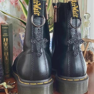 May include: A pair of black leather ankle boots featuring yellow stitching along the sole, a side zipper, and a black heel pull tab with yellow AirWair text. A silver-toned metal ring hangs from each pull tab, holding a black ornate cross charm.