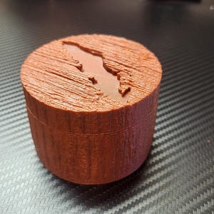 Può includere: Una piccola scatola rotonda di legno con un design intagliato di uno stato o un'isola. La scatola è di colore marrone rossiccio e presenta una venatura del legno.
