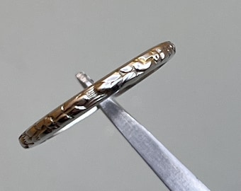 May include: A silver ring with a detailed floral pattern. The ring is held by a silver tool, showcasing the intricate design. The ring's surface reflects light, highlighting the embossed leaves and vines.