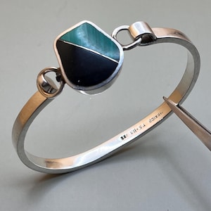 May include: A silver-toned bangle bracelet with a geometric centerpiece. The centerpiece features a split design with a black and green stone. The bracelet has a hook closure and is marked "MEXICO T.S-B3".
