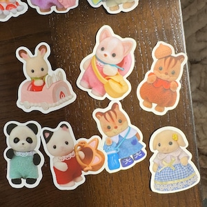 May include: A selection of colourful stickers depicting animal characters in outfits. The stickers include a panda, a cat with a watering can, a rabbit in a car, and other animal figures. The stickers have a white border.