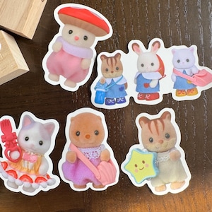 May include: A selection of colourful stickers depicting animal characters in various outfits. The stickers include a mushroom character, a cat in a strawberry dress, and other animal figures holding accessories like a star and a bag.