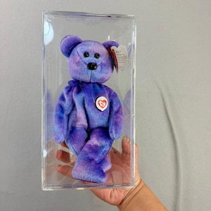 May include: A purple and blue tie-dye plush teddy bear with black button eyes and a black nose. The bear features a red and white heart-shaped tag on its chest and a small tag attached to its ear. It is enclosed in a clear rectangular plastic display case.