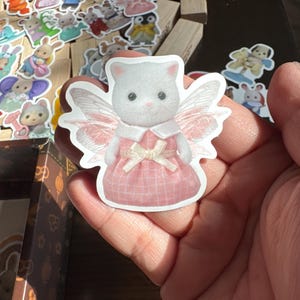 May include: A white cat sticker with pink fairy wings and a pink plaid dress. The sticker is held in a hand, with other animal stickers and wooden blocks in the background. The cat has a white bow and a pink dress.