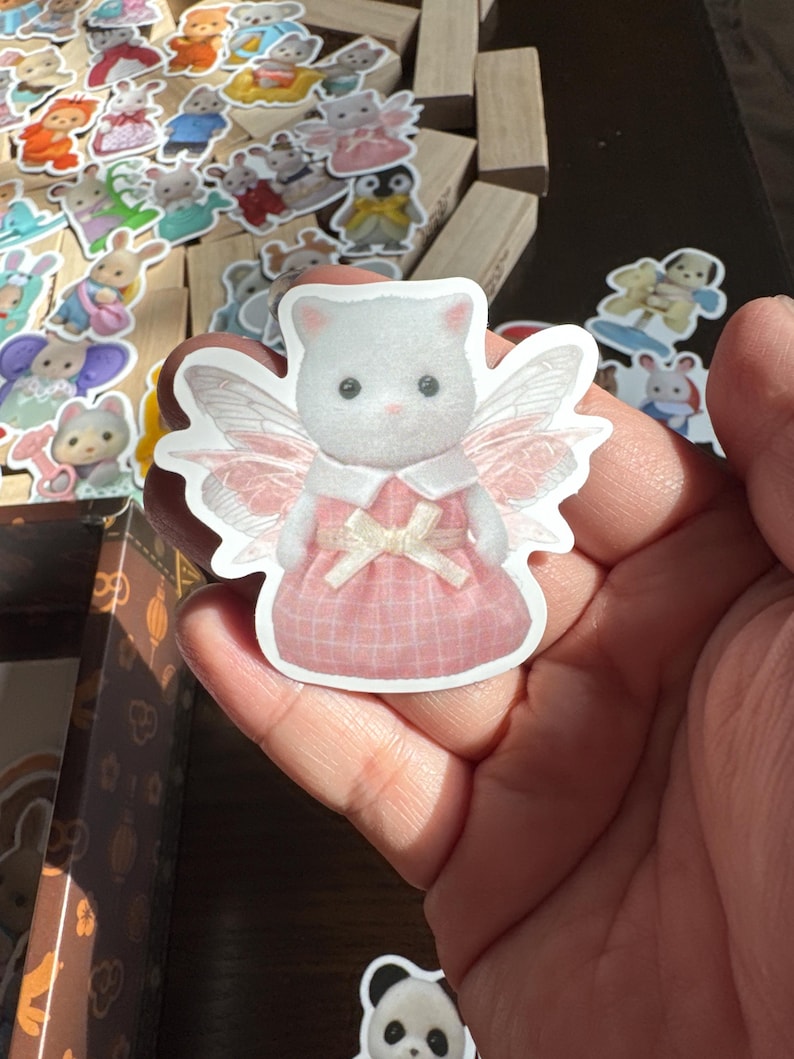 May include: A white cat sticker with pink fairy wings and a pink plaid dress. The sticker is held in a hand, with other animal stickers and wooden blocks in the background. The cat has a white bow and a pink dress.