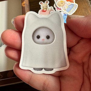 May include: A sticker of a white cat-shaped ghost costume with a round face opening. The cat has a fluffy white face with black eyes and a pink nose. Other stickers of animal characters are in the background.