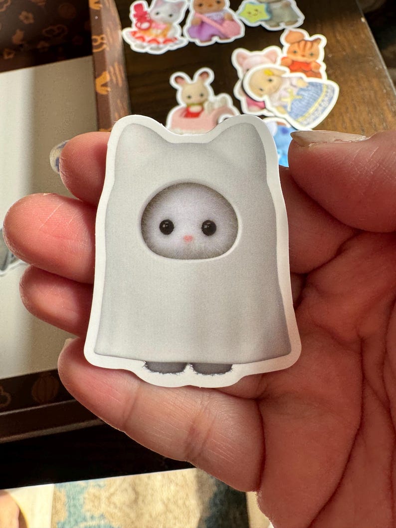 May include: A sticker of a white cat-shaped ghost costume with a round face opening. The cat has a fluffy white face with black eyes and a pink nose. Other stickers of animal characters are in the background.