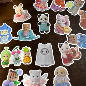 May include: A selection of colourful die-cut stickers showcasing animal characters in playful attire. The stickers include a panda, a bunny in a car, a bear on a rocking horse, and a cat in a dress. The stickers are on a brown surface.
