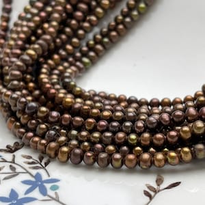 Puede incluir: Primer plano de varios hilos de pequeñas perlas de color marrón oscuro con matices verdes y dorados iridiscentes. Las perlas redondas están ensartadas, creando una apariencia texturizada y elegante. Ideal para la creación de joyas.