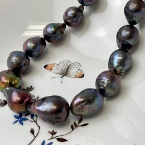 Puede incluir: Un collar de grandes perlas grises oscuras iridiscentes. Las perlas tienen forma de lágrima y están ensartadas. El collar descansa sobre un plato blanco con un diseño de mariposa y flores.