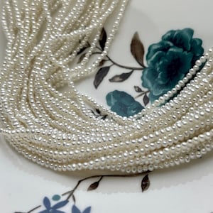 May include: A strand of white freshwater pearls, arranged in a pile on a white surface with a floral pattern.