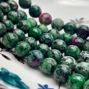 Puede incluir: Primer plano de una colección de cuentas de piedras preciosas facetadas. Las cuentas son principalmente verdes con inclusiones rojas y rosas. Las cuentas están dispuestas sobre una superficie blanca con un diseño floral.