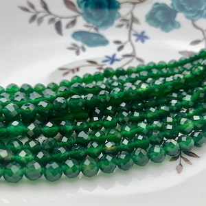Puede incluir: Una hebra de cuentas de piedras preciosas facetadas de color verde oscuro. Las cuentas son redondas y tienen un acabado liso y pulido.