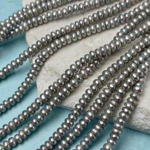 May include: Close-up of multiple strands of gray pearl beads. The beads are small, round, and evenly sized, creating a smooth, lustrous surface. The strands are arranged in a layered pattern, suggesting a necklace or jewelry design.