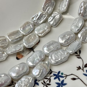 May include: Close-up of strands of white, irregularly shaped freshwater pearls. The pearls have a lustrous, pearlescent sheen. The background is a white plate with a floral design. These pearls are ideal for jewelry making.