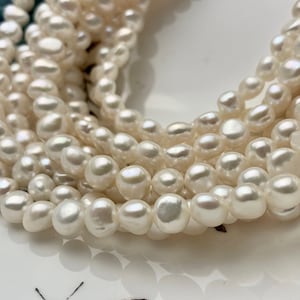 Puede incluir: Una hebra de perlas de agua dulce blancas, con una forma ligeramente irregular, dispuestas en un montón suelto sobre una superficie blanca.