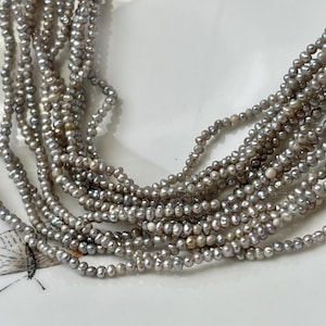 May include: Strands of gray freshwater pearls cascade across a white surface. The pearls are small and irregularly shaped, creating a delicate and elegant appearance. The pearls are arranged in multiple strands.