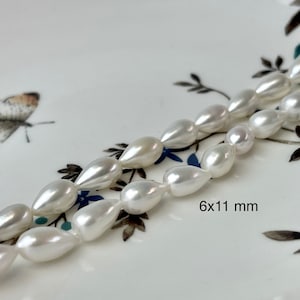 May include: A strand of white pearlescent teardrop-shaped beads on a white background with a floral pattern. The beads are approximately 6x11 mm in size.