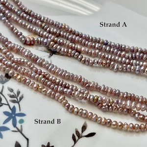 May include: Close-up of multiple strands of pinkish-purple freshwater pearls. The strands are labeled "Strand A" and "Strand B". The pearls have an irregular shape and a subtle luster, suggesting they are natural. The background is a white surface with a floral design.