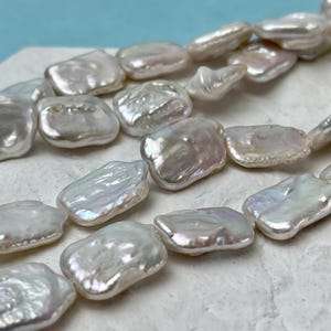May include: Strands of lustrous, irregular-shaped, white freshwater pearls. The pearls have a natural, iridescent sheen, reflecting light with subtle pink and silver hues. These organic-shaped pearls are strung together, showcasing their unique form.