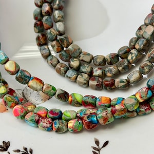 May include: Strands of colorful gemstone beads in shades of green, blue, red, and brown. The beads are square-shaped and strung together, creating a textured and vibrant appearance. The beads are displayed on a white surface.