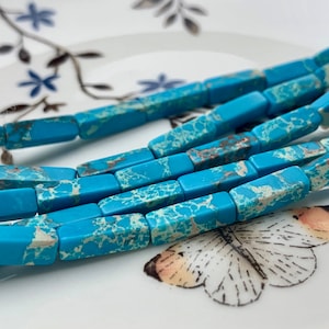 May include: Strands of turquoise rectangular beads with a marbled pattern. The beads are a vibrant blue with brown and white veining. The beads are displayed on a white plate with a floral design.