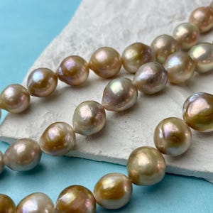 May include: Close-up of several strands of lustrous, irregularly shaped pearls. The pearls display a range of colours, including gold, cream, and hints of pink, resting on a white, textured surface against a light blue backdrop.
