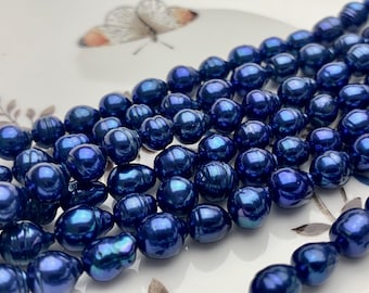 Puede incluir: Primer plano de una colección de perlas barrocas azul oscuro. Las perlas tienen un brillo lustroso e iridiscente, que refleja tonos de azul y púrpura. Las formas irregulares de las perlas son visibles, lo que sugiere un origen natural. Las perlas están dispuestas sobre una superficie blanca.