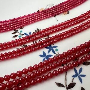 May include: A strand of red gemstone beads, with multiple strands layered on top of each other. The beads are round and smooth, with a glossy finish.