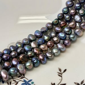 May include: Strands of iridescent, baroque-shaped pearls in shades of deep blue, purple, and bronze. The pearls are arranged in three parallel rows, showcasing their unique, irregular shapes and lustrous surfaces. The pearls are displayed on a white surface.