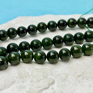 Puede incluir: Primer plano de cuentas de piedras preciosas pulidas de color verde oscuro. Las cuentas son redondas y tienen un acabado brillante, que refleja la luz. Las cuentas están ensartadas, listas para la fabricación de joyas. Fondo claro y texturizado con un fondo azul.