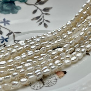 Könnte beinhalten: Eine Strähne weißer Süßwasserperlen, jede Perle hat eine einzigartige, unregelmäßige Form. Die Perlen sind in einem lockeren, fließenden Muster angeordnet, was einen zarten und eleganten Look erzeugt.