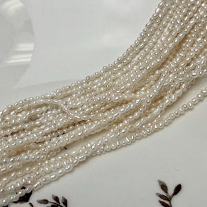 May include: Close-up of multiple strands of creamy white freshwater pearls. The pearls are small, oval-shaped, and strung together, creating a flowing, textured appearance. The background is a white surface.