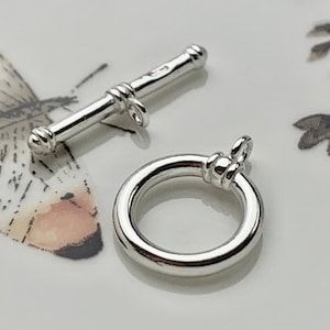 Könnte beinhalten: Zwei silberfarbene Metallverschlüsse zum Schmuckbasteln. Ein Verschluss ist ein Toggle-Verschluss mit rundem Ende. Der andere Verschluss ist ein runder Ring.