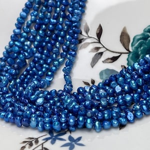 May include: A strand of blue freshwater pearls, with a slightly irregular shape, arranged in a loose pile on a white surface with a floral pattern.