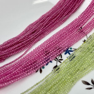 May include: Two strands of gemstone beads, one pink and one green, are laid out on a white surface. The beads are small and round, and they have a slightly faceted appearance.