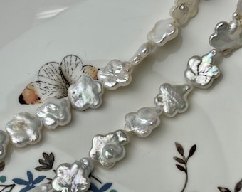 Peut inclure: Deux rangées de perles blanches de forme irrégulière, ressemblant à des pétales de fleurs. Les perles ont un éclat brillant et sont enfilées ensemble. Le fond est une assiette blanche avec un motif floral, ajoutant un contraste délicat aux bijoux.