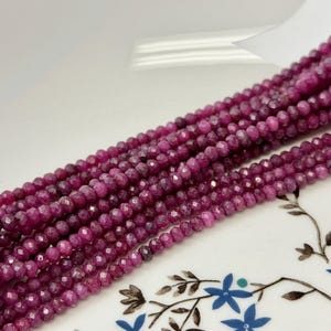 May include: A long strand of small, faceted, deep magenta-colored beads arranged in multiple parallel rows. The beads have a reflective, textured surface and are strung together, creating a dense, uniform appearance across the entire length.