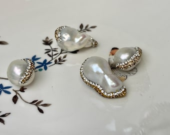 May include: Four irregularly shaped, white pearl beads with a gold and clear crystal border. The pearls are arranged on a white floral patterned surface. The pearls have a lustrous, iridescent sheen.