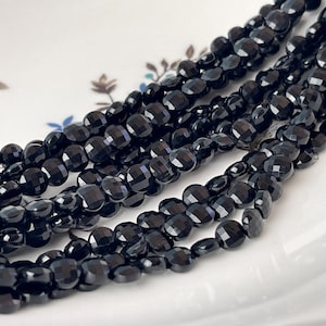 May include: Close-up of several strands of faceted black gemstone beads. The beads are a deep, glossy black and have a geometric, multi-faceted cut. The beads are arranged on a white surface with a floral design.
