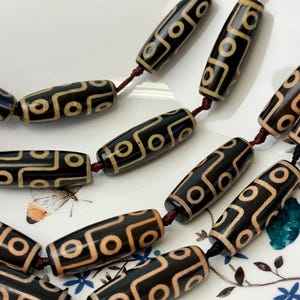 Może przedstawiać: Naszyjnik z czarno-beżowych koralików w kształcie beczułek z geometrycznymi wzorami. Koraliki są nawleczone na ciemnoczerwony sznurek i prezentowane na białym talerzu z kwiatowymi detalami. Koraliki mają błyszczące wykończenie.